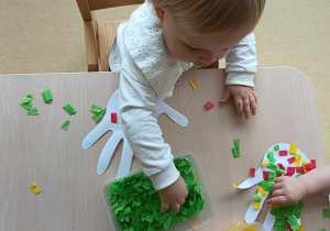 Dziewczynka wybiera z plastikowego prostokątnego pojemnika pocięta zielona bibułę.