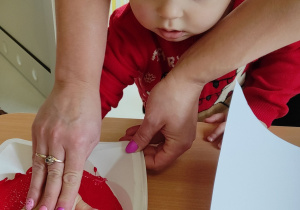 Jedna z opiekunek pomaga Szymonkowi odcisnąć dłoń w farbie znajdującej się na plastikowym talerzyku.